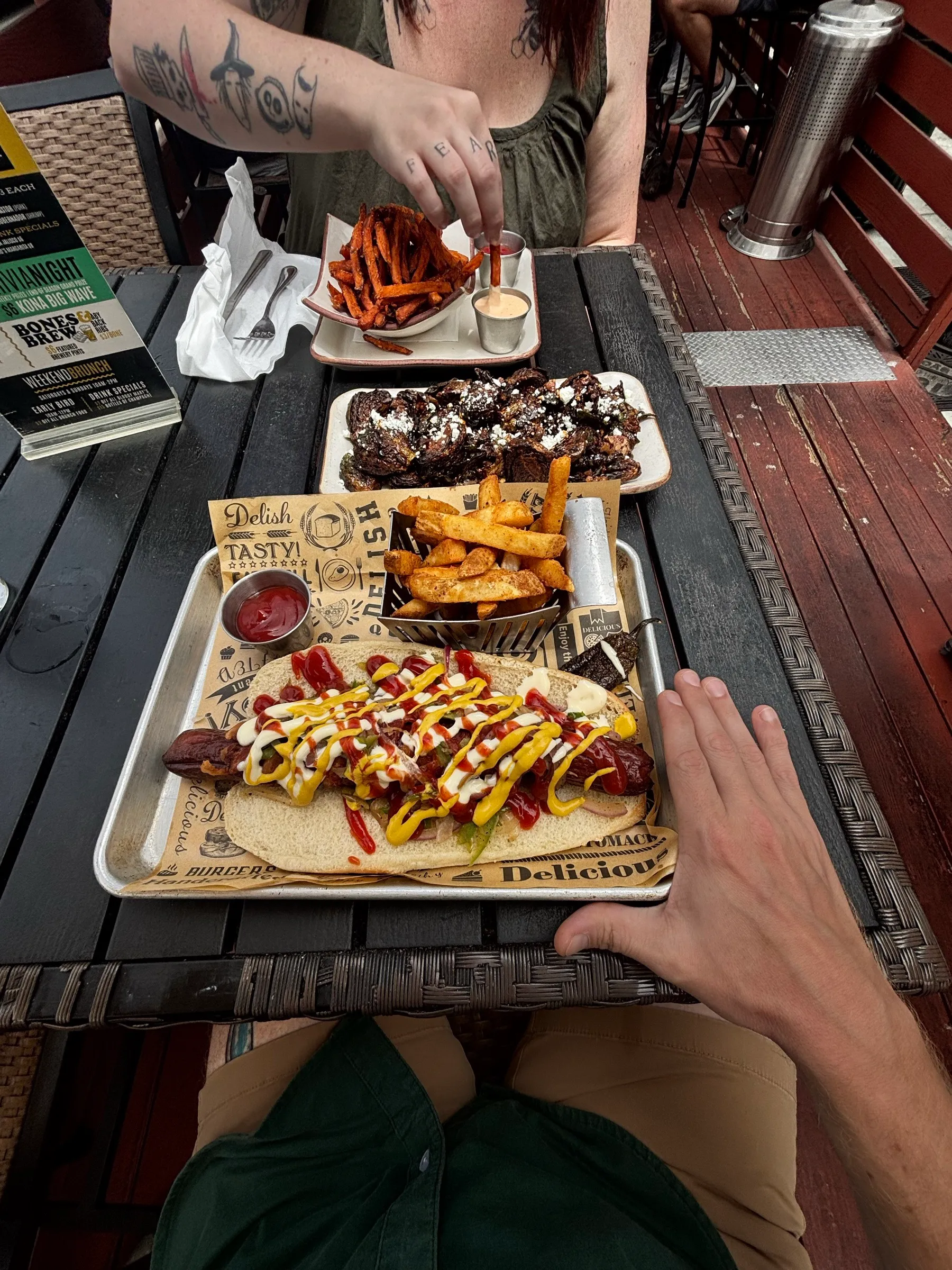 A bacon-wrapped hot dog zigzagged with condiments, fries, and brussels sprouts on a patio table